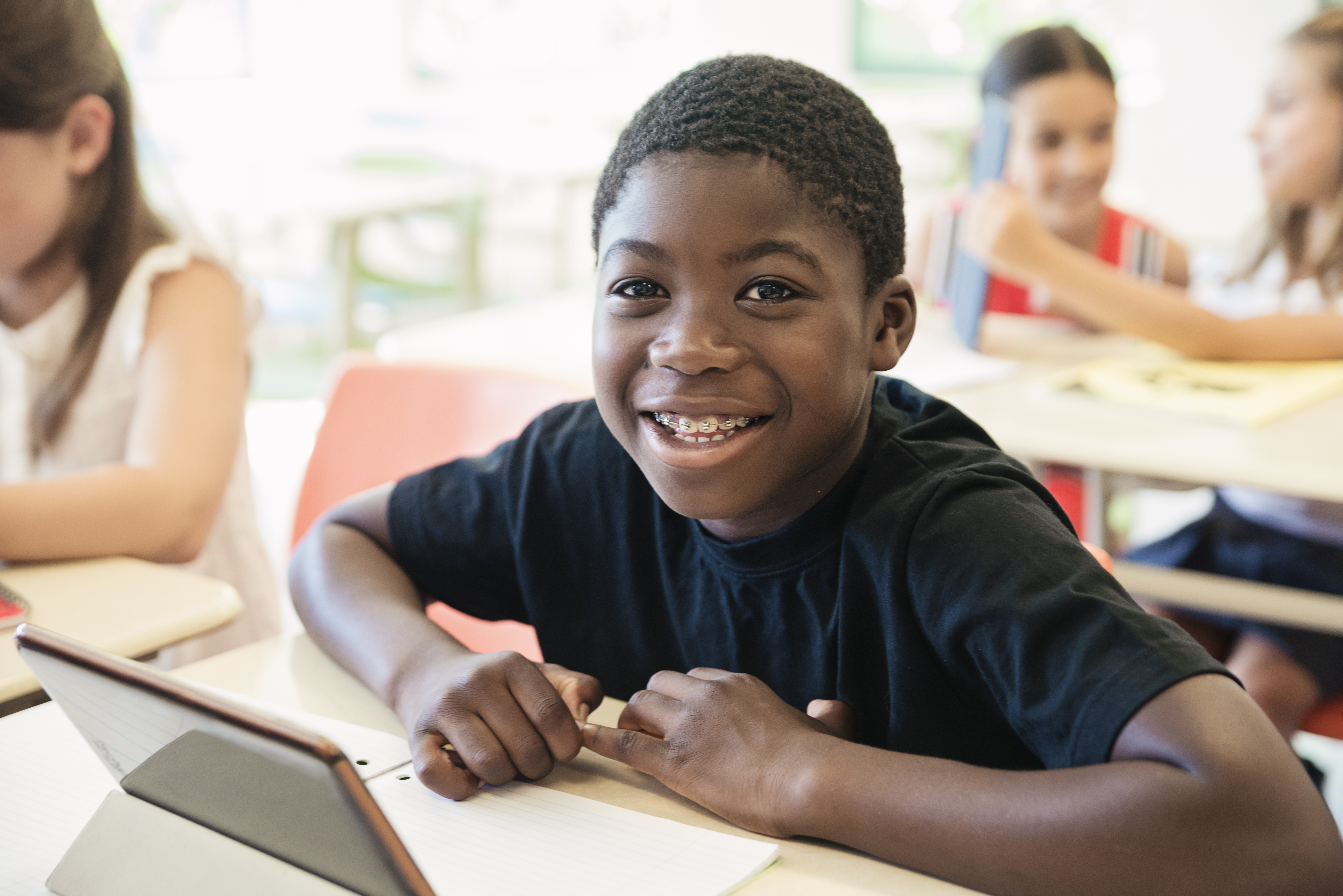 African-american boy using electronic tablet in classroom. He wear braces and is looking at the camera with a smile. Some schoolgirls in the background. Horizontal waist up indoors shot with copy space. This was taken in Quebec, Canada.